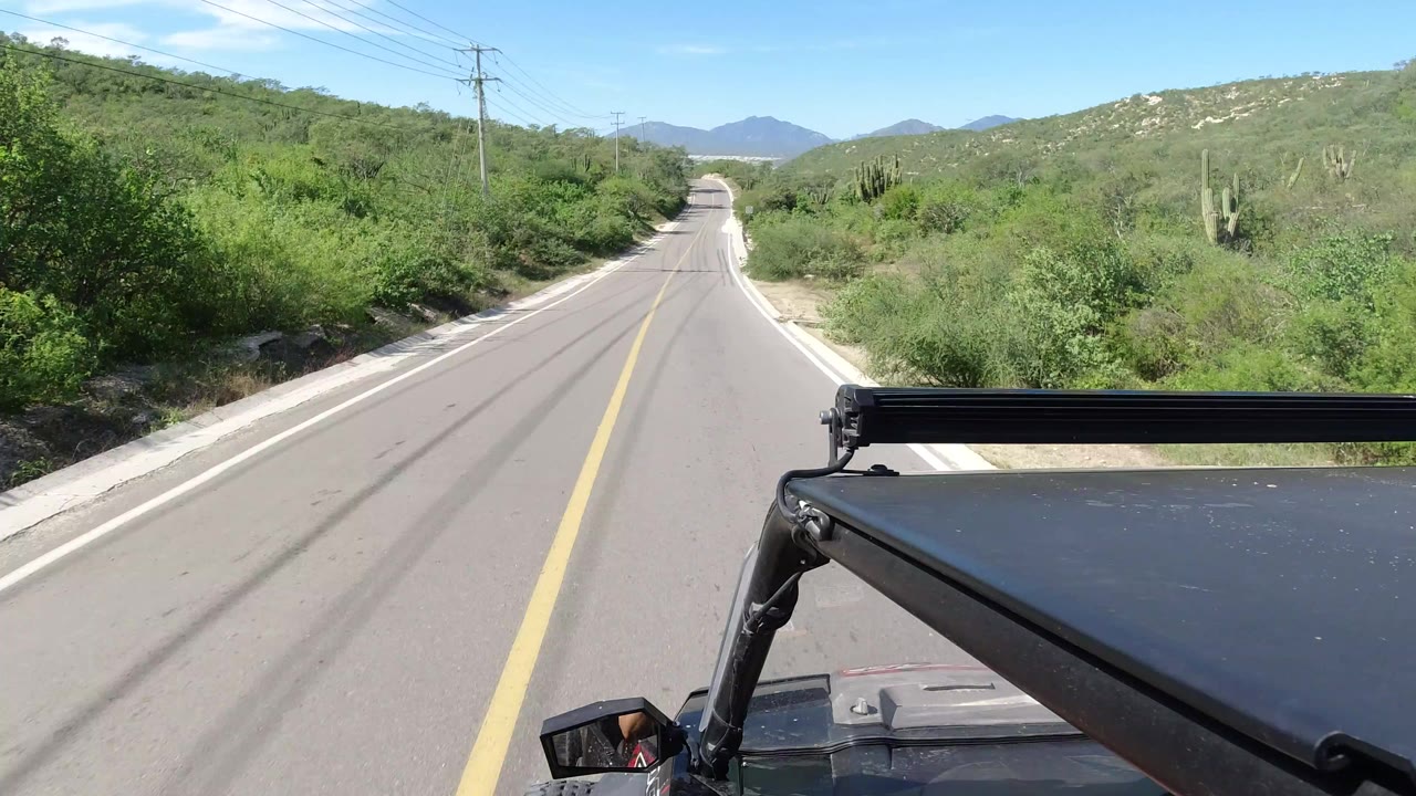 Opening Los Cabos road view from a vehicle, with a yellow centerline, dense green roadside shrubs, distant hills, blue sky, and no visible people.