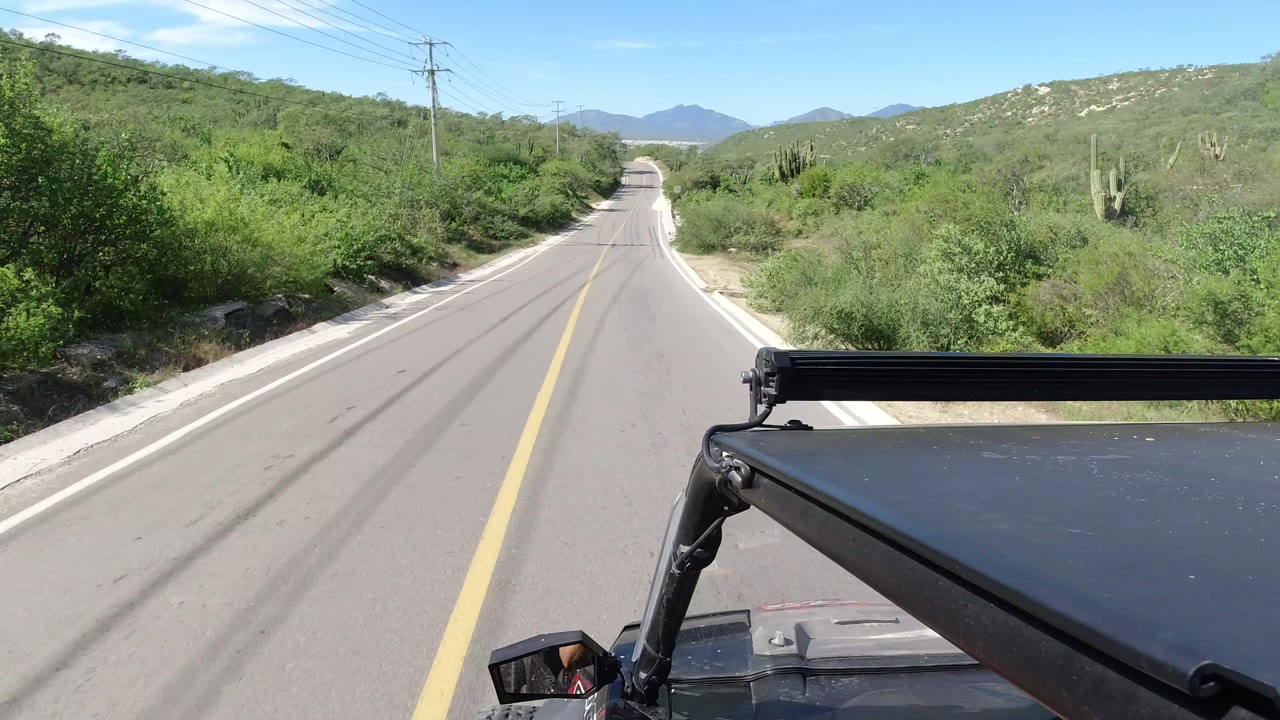Los Cabos vehicle-mounted road perspective continues, with the lane centered between green brush, pale shoulders, utility poles, and no visible people.