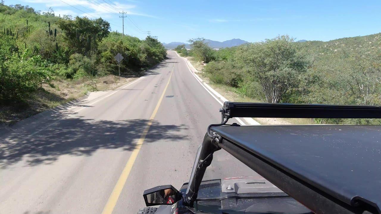 Forward-facing vehicle POV on a paved road in Los Cabos, Mexico, with green brush on both sides, distant hills, blue sky, and no clear faces visible.