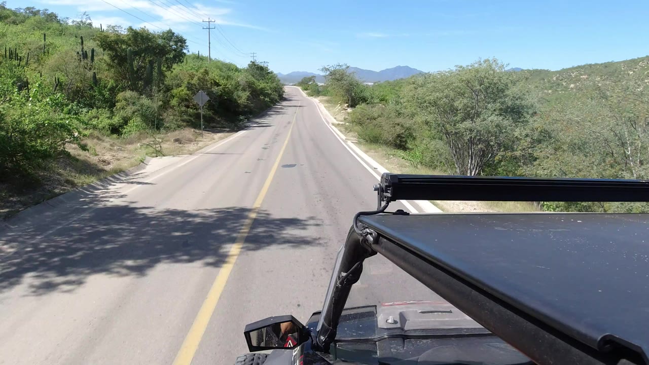 Vehicle-mounted road view in Los Cabos, Mexico, with the black roof panel in the lower frame, a yellow center line, dense green shrubs, and no clear faces visible.