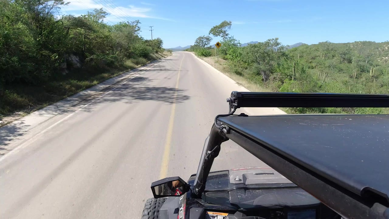 Opening view of a Los Cabos, Mexico road bordered by green brush and low hills, with a black vehicle roof bar in the lower right and no recognizable people visible.