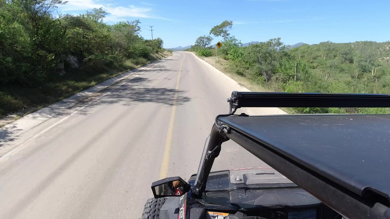 A Los Cabos, Mexico roadway stretches ahead under bright sun, with scrubby vegetation on both sides, distant hills, and the vehicle frame fixed along the lower right edge.