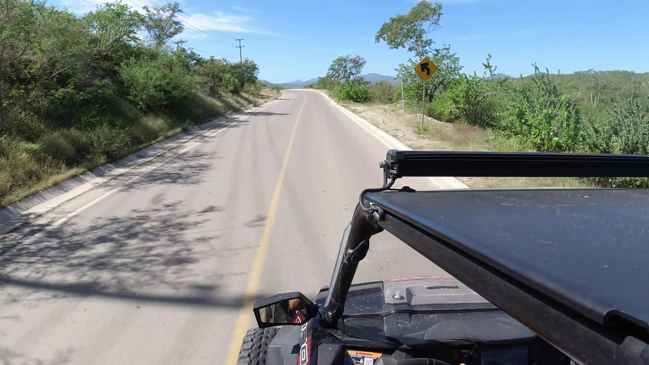The road begins to bend through Los Cabos, Mexico brushland, with a yellow curve warning sign on the right shoulder and no recognizable people visible.