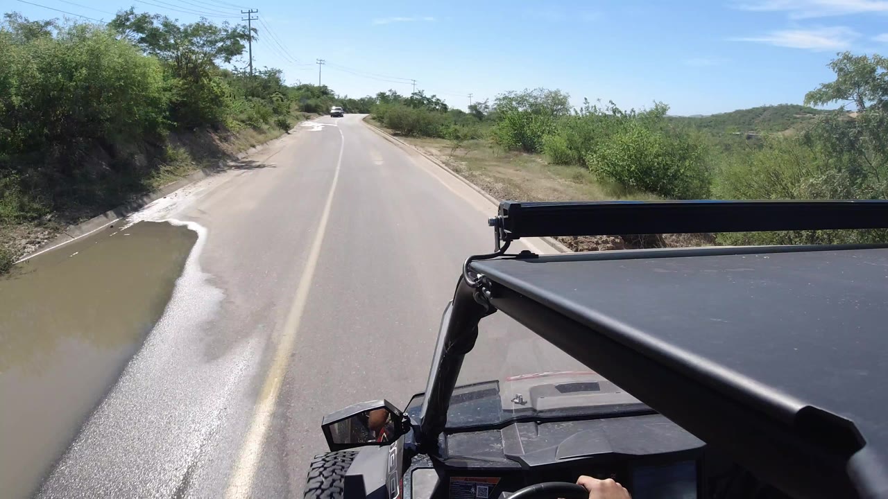 Opening view in Los Cabos, Mexico, from an open vehicle on a paved road, with a muddy roadside channel on the left, green brush on both sides, blue sky above, and no visible people.