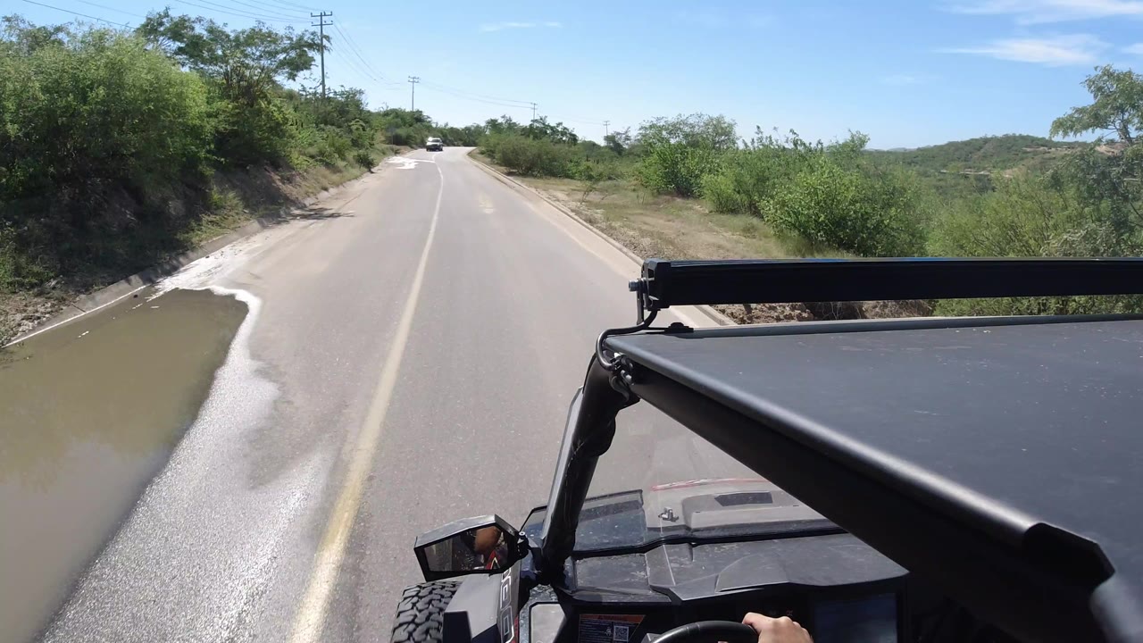 Second frame in Los Cabos, Mexico, keeps the open vehicle roof in the foreground as the paved road continues beside a muddy shoulder channel, roadside shrubs, utility poles, and no visible people.