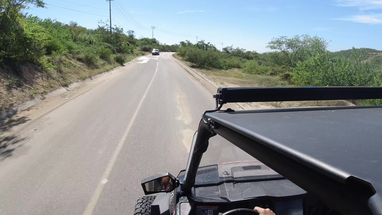 Vehicle-mounted view of a road cresting ahead with scrub vegetation and utility poles.