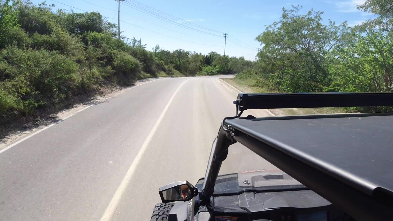 Start of a Los Cabos road clip showing a pale paved lane curving left, green roadside brush, a black vehicle hood in the foreground, and no visible people.