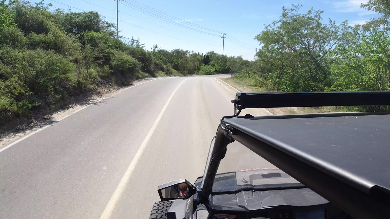 Los Cabos roadway continues past dense green brush, with the black vehicle hood and roof bar filling the lower right foreground and no visible people.