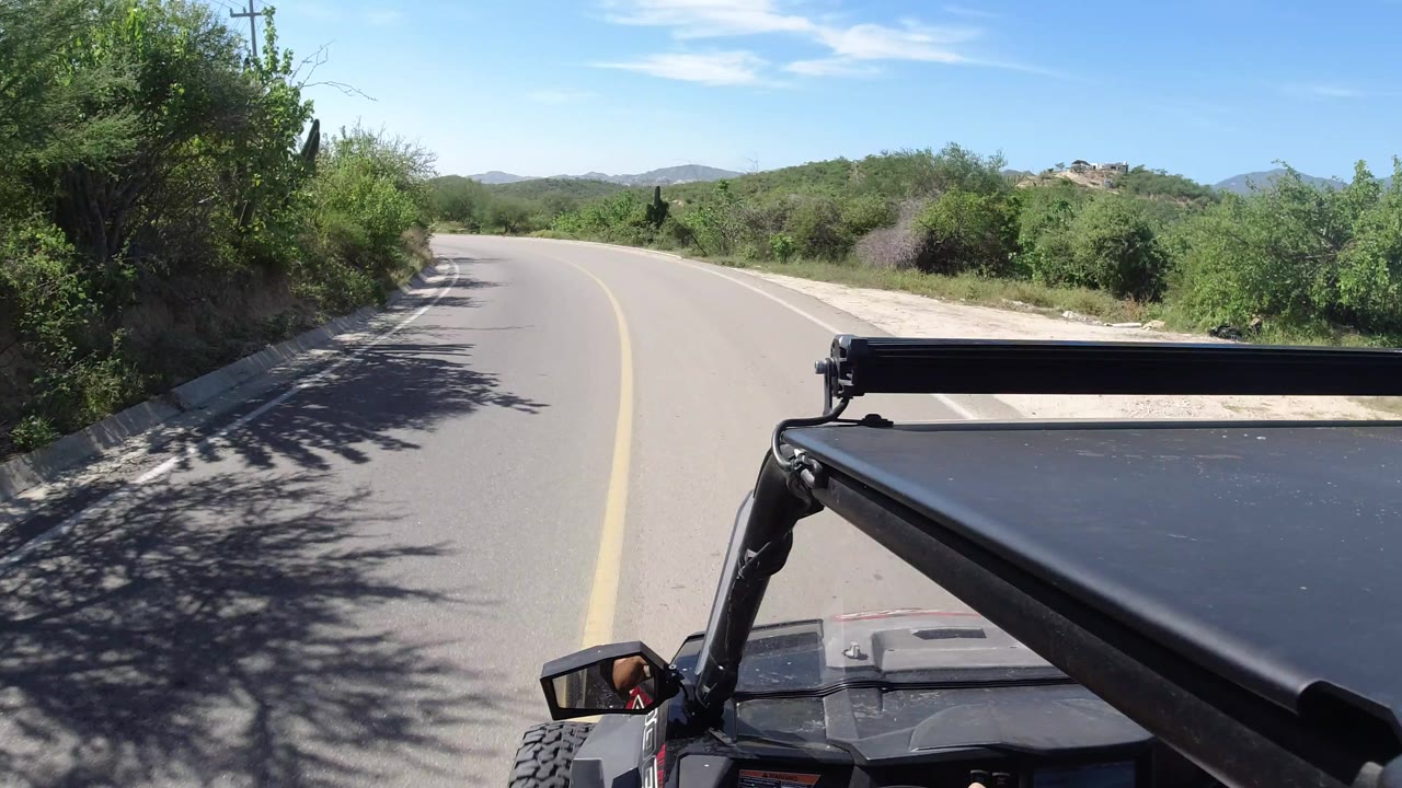 A Los Cabos paved road bends left past thick vegetation, with the black roof support of the vehicle filling the lower right foreground.