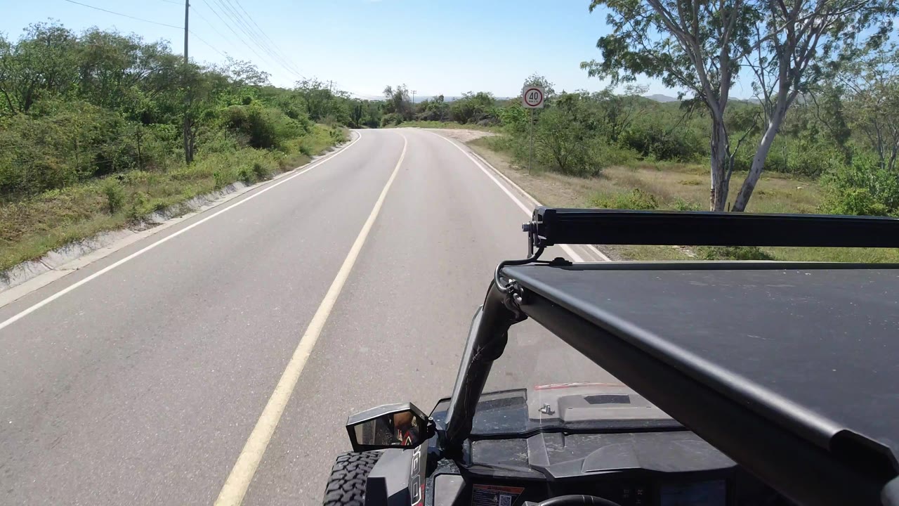 A Los Cabos road continues ahead under bright sun, with the vehicle roof fixed in the lower frame and no visible people.