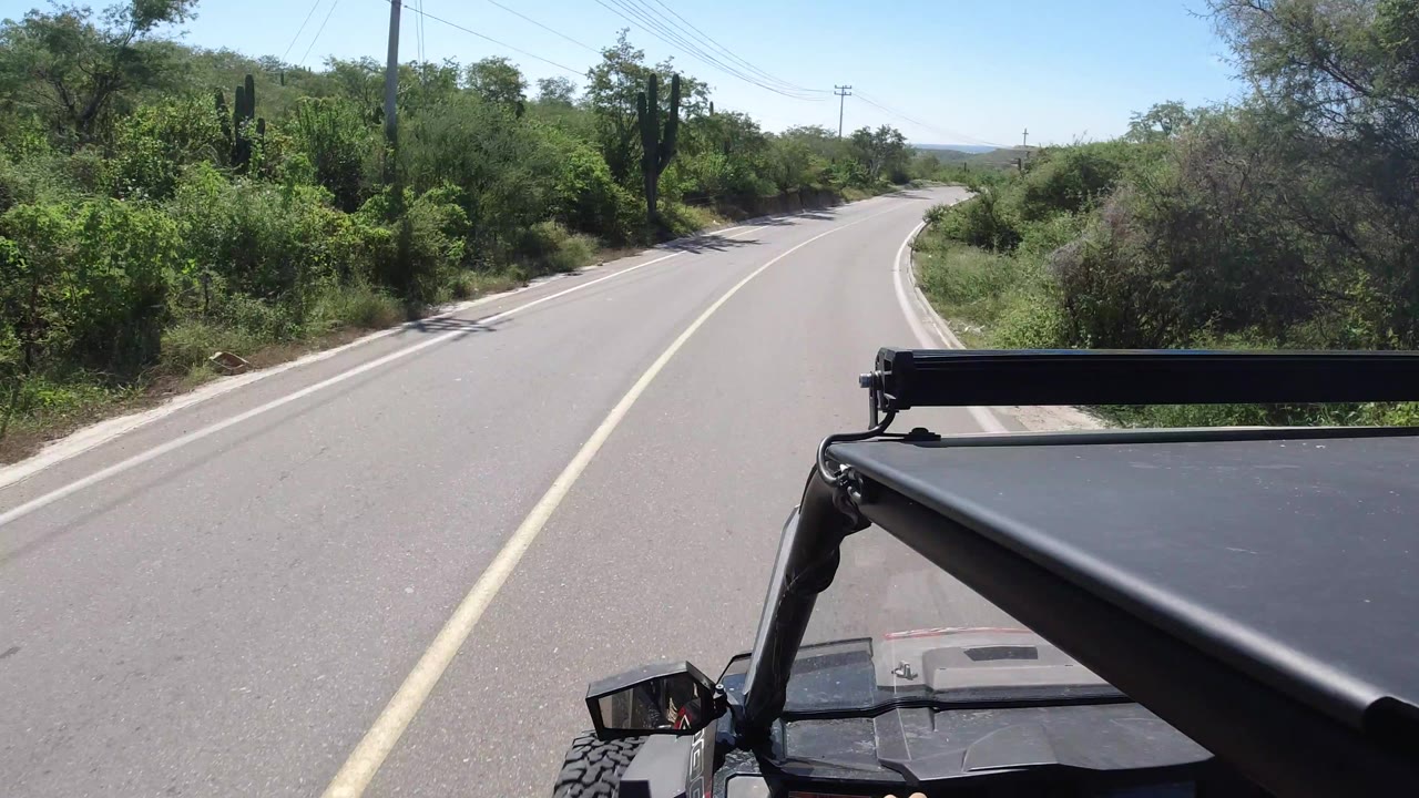 At 0:01, the Los Cabos road continues ahead with the jeep hood and roof bar fixed in the lower frame, dense green brush on both sides, and no visible people.