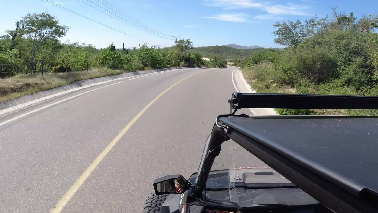 A Los Cabos road view from a vehicle roof area shows the black roof rack and light bar in the foreground, a yellow center line, green roadside brush, distant hills, blue sky, and no visible people.