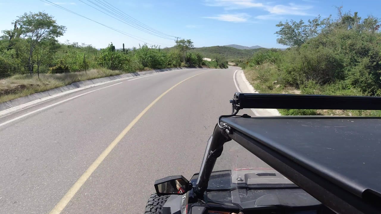 A Los Cabos driving POV shows the road beginning to bend, with the vehicle light bar across the lower frame, dense green shrubs along the shoulder, low hills ahead, and no visible people.