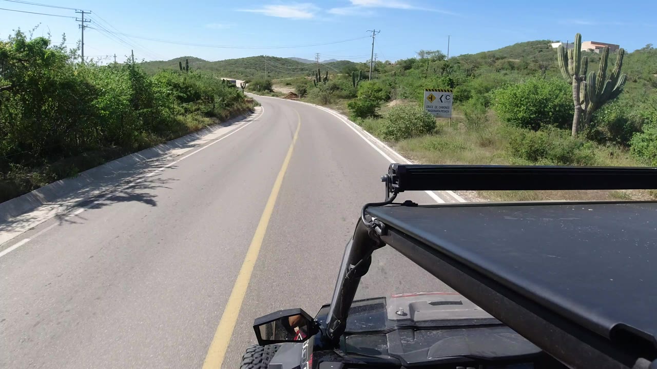 The Wrangler continues along a narrow paved road in Los Cabos, Mexico, passing a small roadside sign, cactus, shrubs, utility poles, and no visible people.