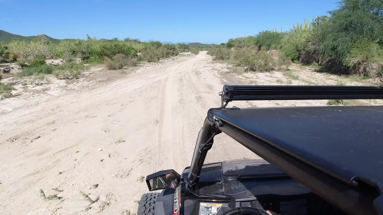 Start of the Los Cabos, Mexico buggy ride with a sandy track ahead, green brush on both sides, distant low hills, and no visible people.