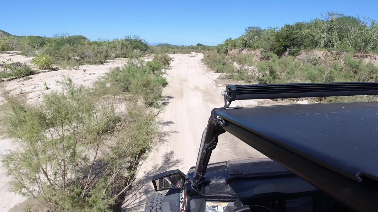 At 0:00 in Los Cabos, Mexico, the Wrangler hood and roof light bar appear at lower right beside a sandy road bordered by green brush, with no visible people.