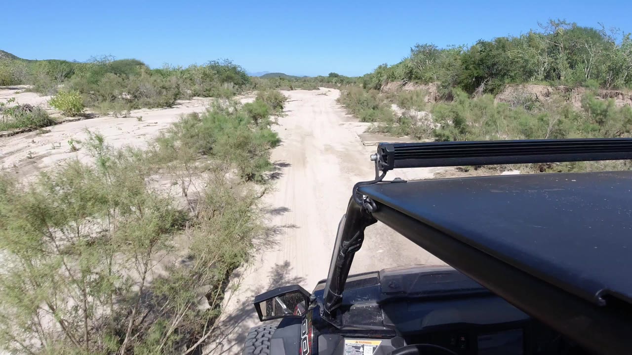 At 0:01 in Los Cabos, Mexico, the sandy track continues ahead past roadside shrubs, with the Wrangler hood and light bar steady in the lower right and no visible people.
