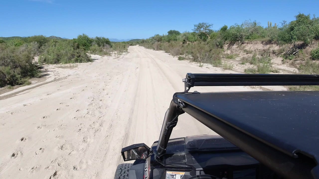 Forward view from a black off-road vehicle on a sandy road in Los Cabos, Mexico, with scrub vegetation on both sides and no visible people.