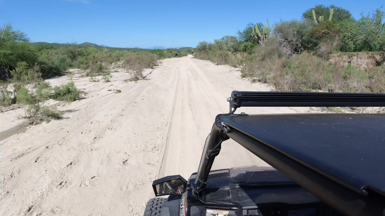 Forward view over a black vehicle hood on a sandy road through Los Cabos desert brush, with low hills and blue sky beyond and no visible people.