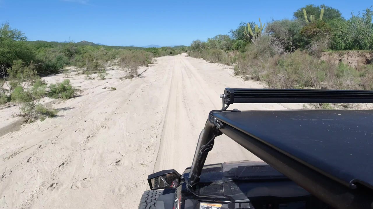 A pale sandy track continues ahead in Los Cabos, Mexico, with scrub and cactus along both sides, distant hills on the horizon, and no visible people.