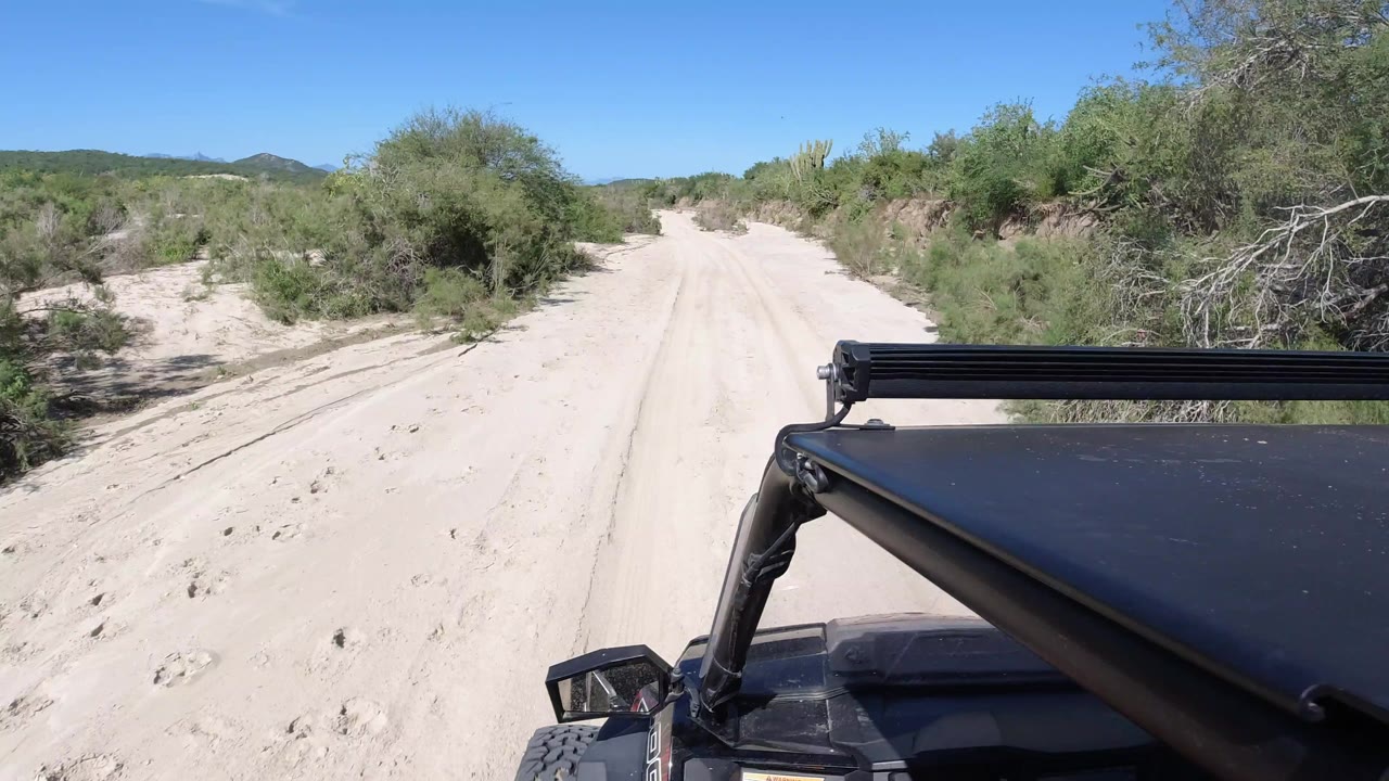 At 0:01, the pale dirt track continues through scrub vegetation in Los Cabos Mexico, with the black vehicle foreground occupying the lower right and no visible people.