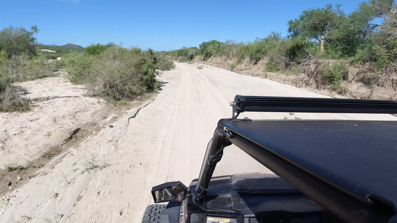 At 0:01 in Los Cabos, Mexico, the sandy road continues through low brush, with the jeep roof fixed in the foreground and a small hillside visible on the horizon with no visible people.
