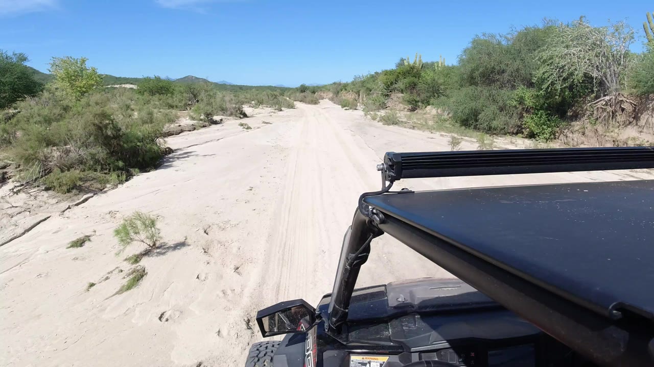 The Los Cabos sandy track continues ahead from the Wrangler viewpoint, with the vehicle roof in the lower right, scrub close to the road edges, low hills beyond, and no visible people.