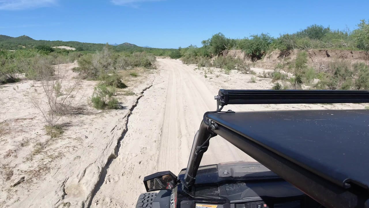 The vehicle-mounted view continues along a pale sandy track in Los Cabos, Mexico, with scrubby green vegetation on both sides and the black roof and light bar filling the lower right foreground.