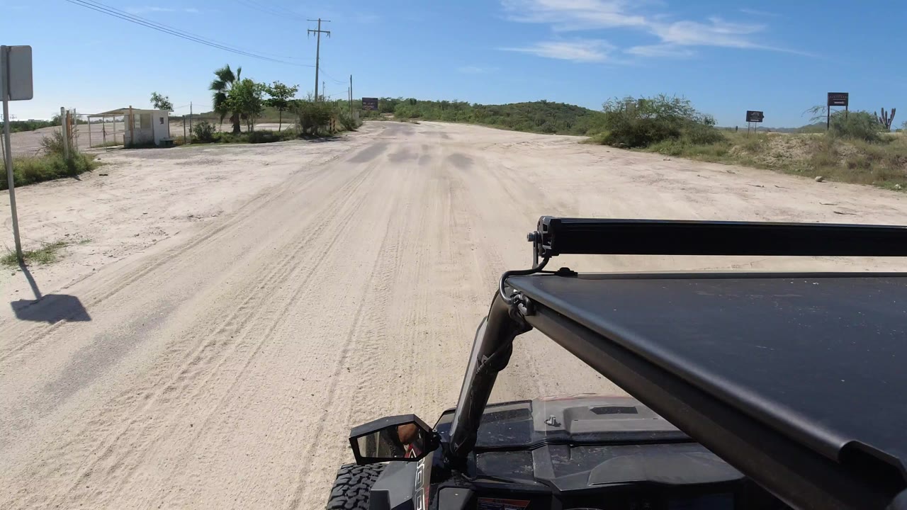 Start of the Los Cabos clip with a black Wrangler roof in the foreground, a pale sandy road ahead, roadside palms and signs, and no visible people.