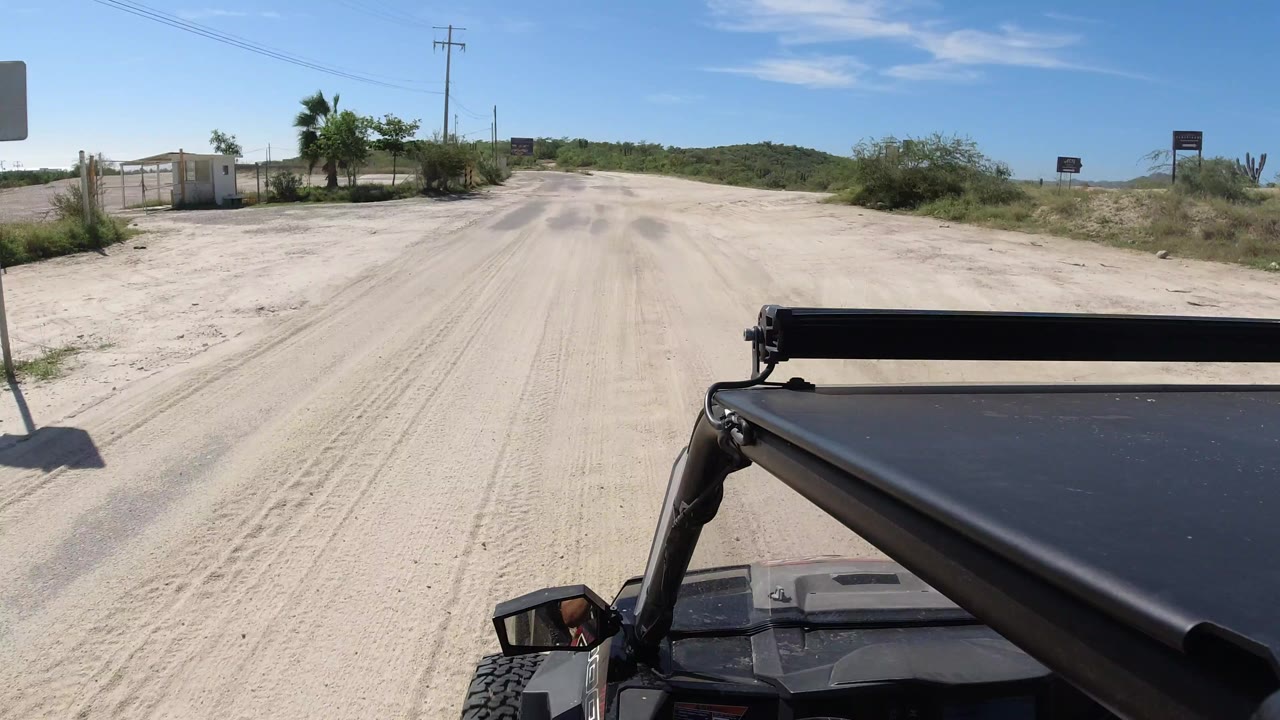 Los Cabos road view from the Wrangler with the vehicle roof steady at the bottom, sand shoulders, utility lines, scrub, low hills, and no visible people.