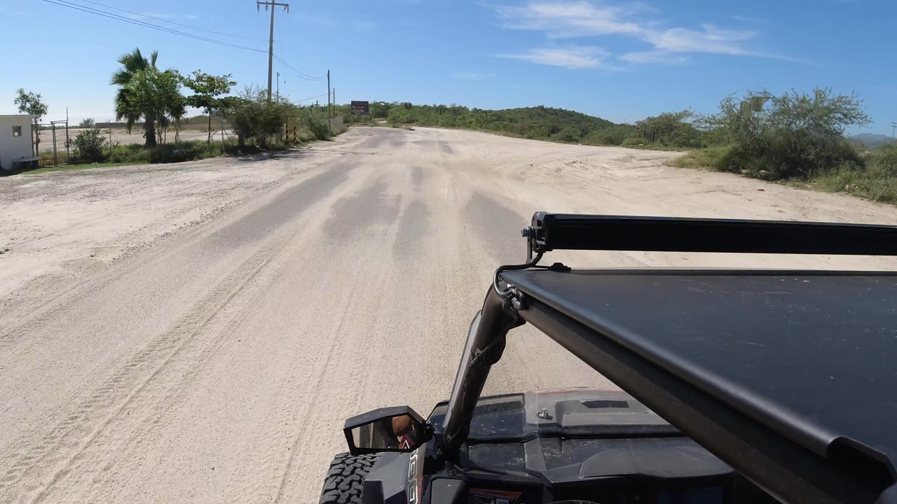 The Los Cabos roadway begins to curve, with the Wrangler foreground, pale road surface, scrubby roadside plants, distant hills, and no visible people.
