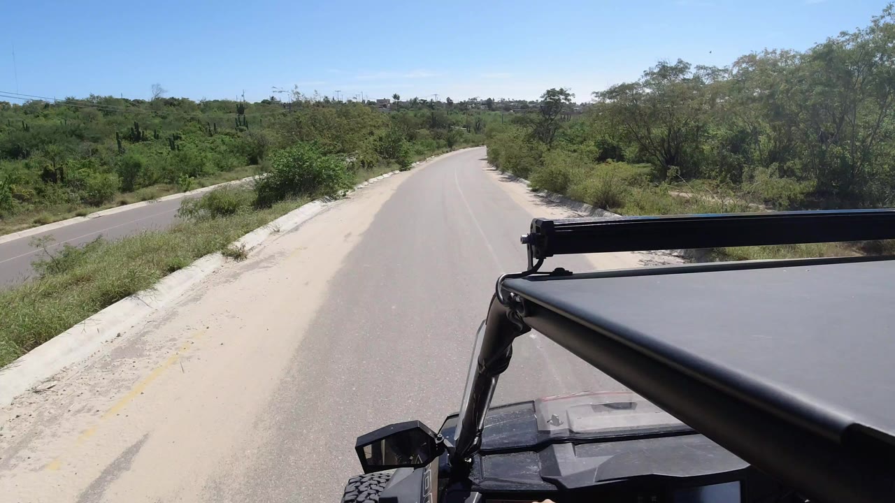 Los Cabos road view from a Wrangler, with the black roof frame in the foreground, a pale sandy shoulder, green brush, and no visible people.
