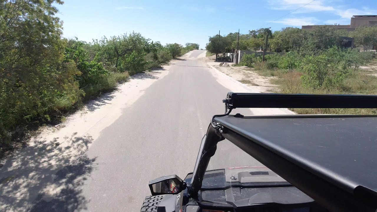 Forward view from an open vehicle on an empty paved road in Los Cabos, Mexico, with green roadside vegetation, sandy shoulders, distant buildings, and no visible people.