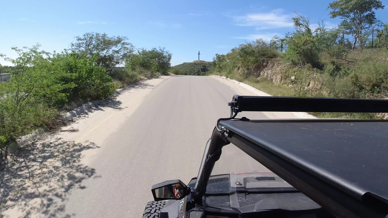Forward vehicle-mounted view along a smooth road in Los Cabos, Mexico, with the Wrangler structure fixed at lower right, roadside brush, sand, distant hill, and no visible people.