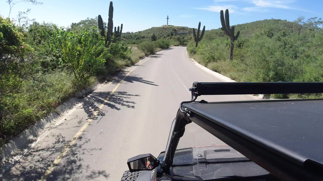 Forward-facing road view in Los Cabos, Mexico with tall cacti on both sides, low green hills ahead, a cross on a distant ridge, and no visible people.