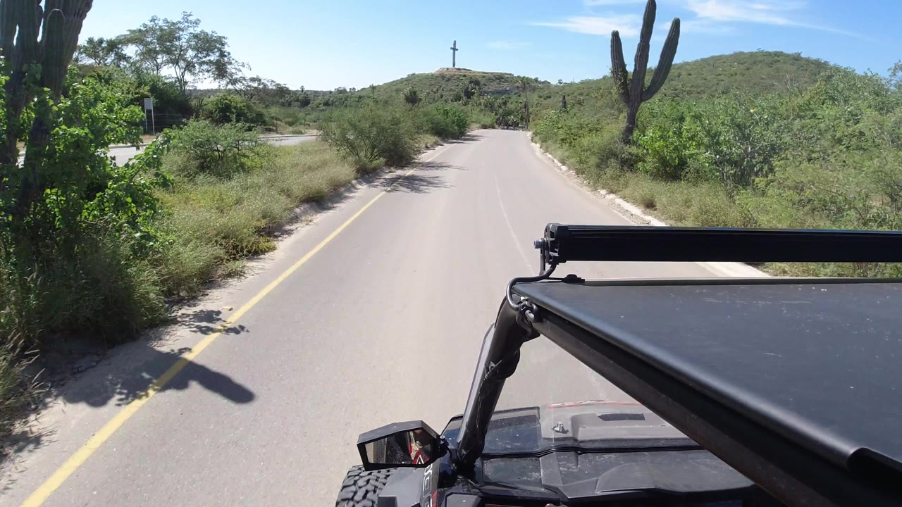 Los Cabos road frame with an open paved lane, cactus shadow across the asphalt, thick green vegetation to the right, low hill ahead, and no visible people.