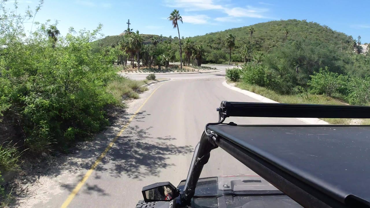 Jeep roof and support bar fill the lower foreground as a curved Los Cabos road leads toward palms, shrubs, a green hillside, blue sky, and no visible people.