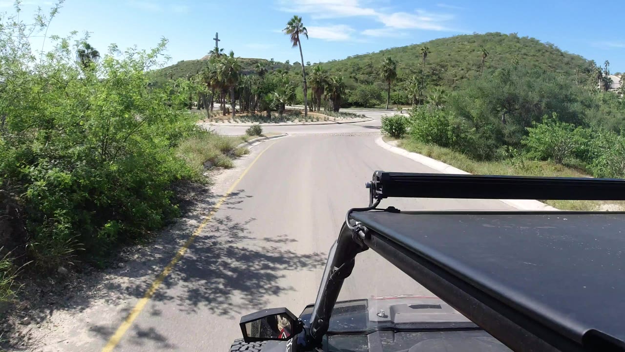 The Jeep foreground remains steady while the road curves left past dense green shrubs, palms, a low hill, bright sky, and no visible people in Los Cabos.
