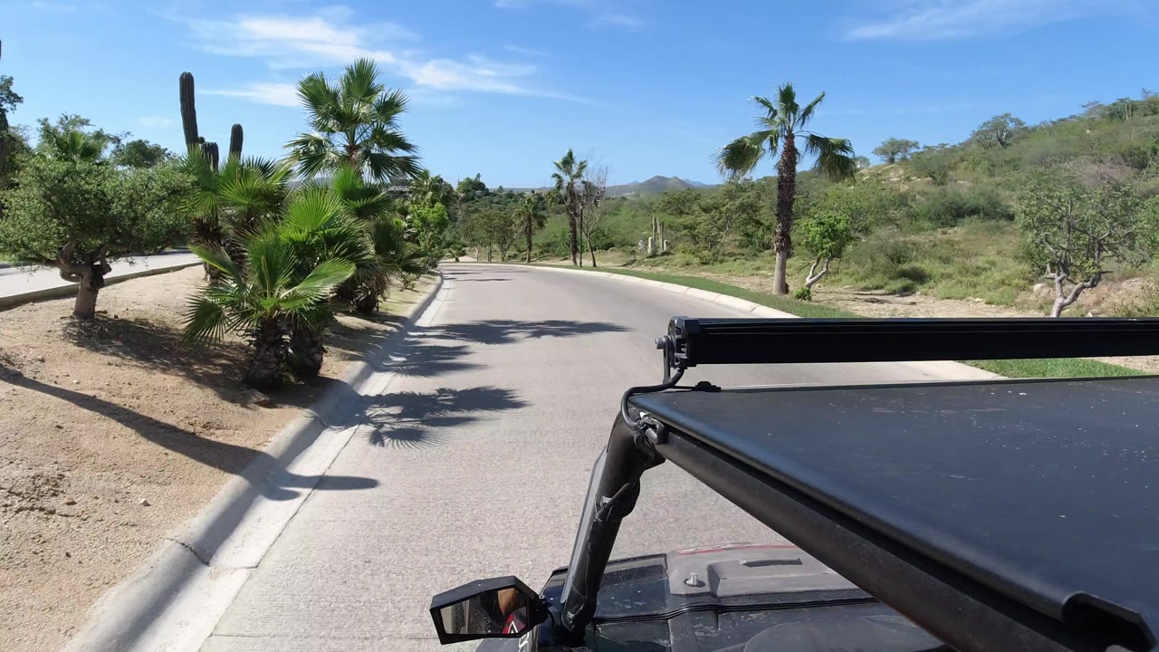 The Los Cabos road view continues with the Jeep roof fixed at the bottom of frame, palm clusters near the curb, dry landscaping, distant hills, and no visible people.