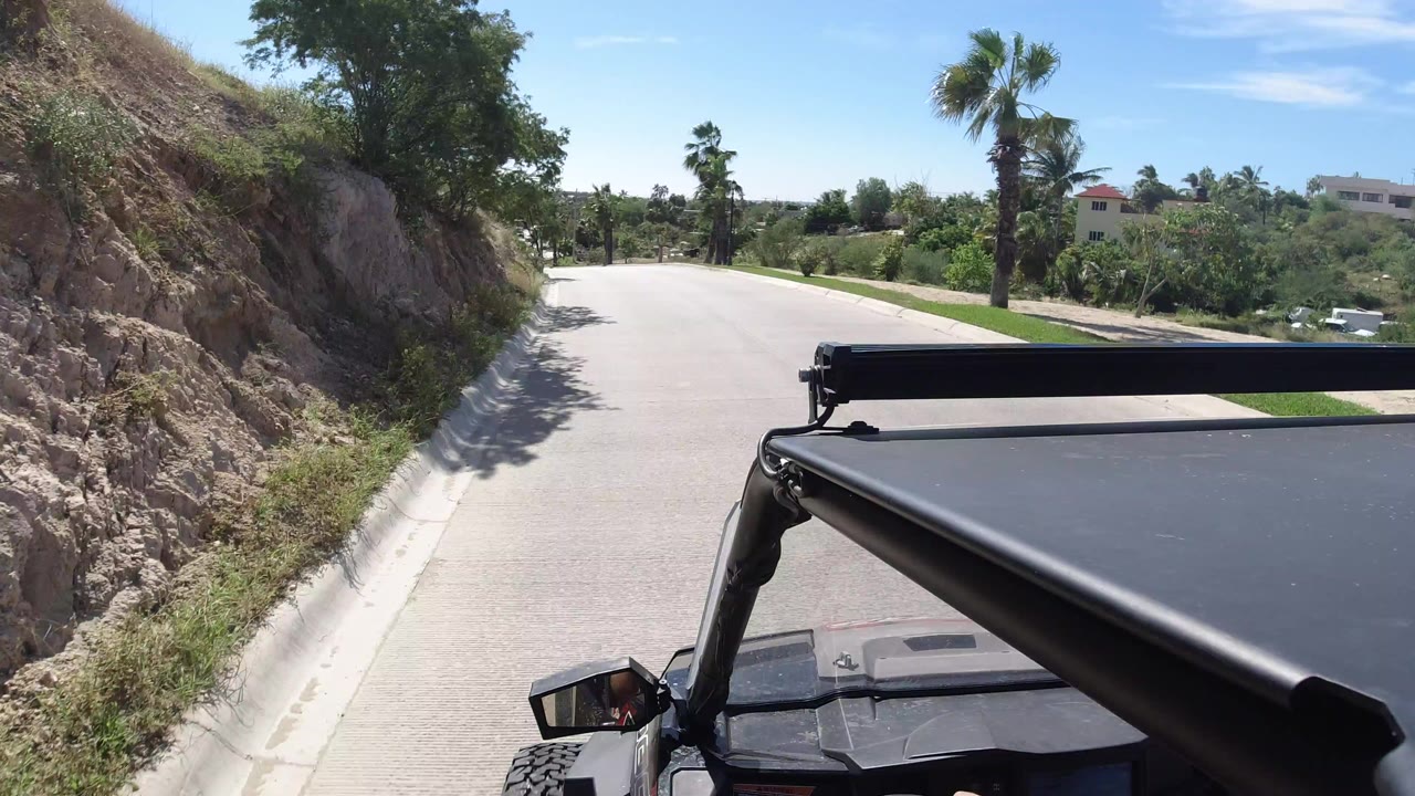 Vehicle roof foreground on a sunny curving road beside a rocky slope.