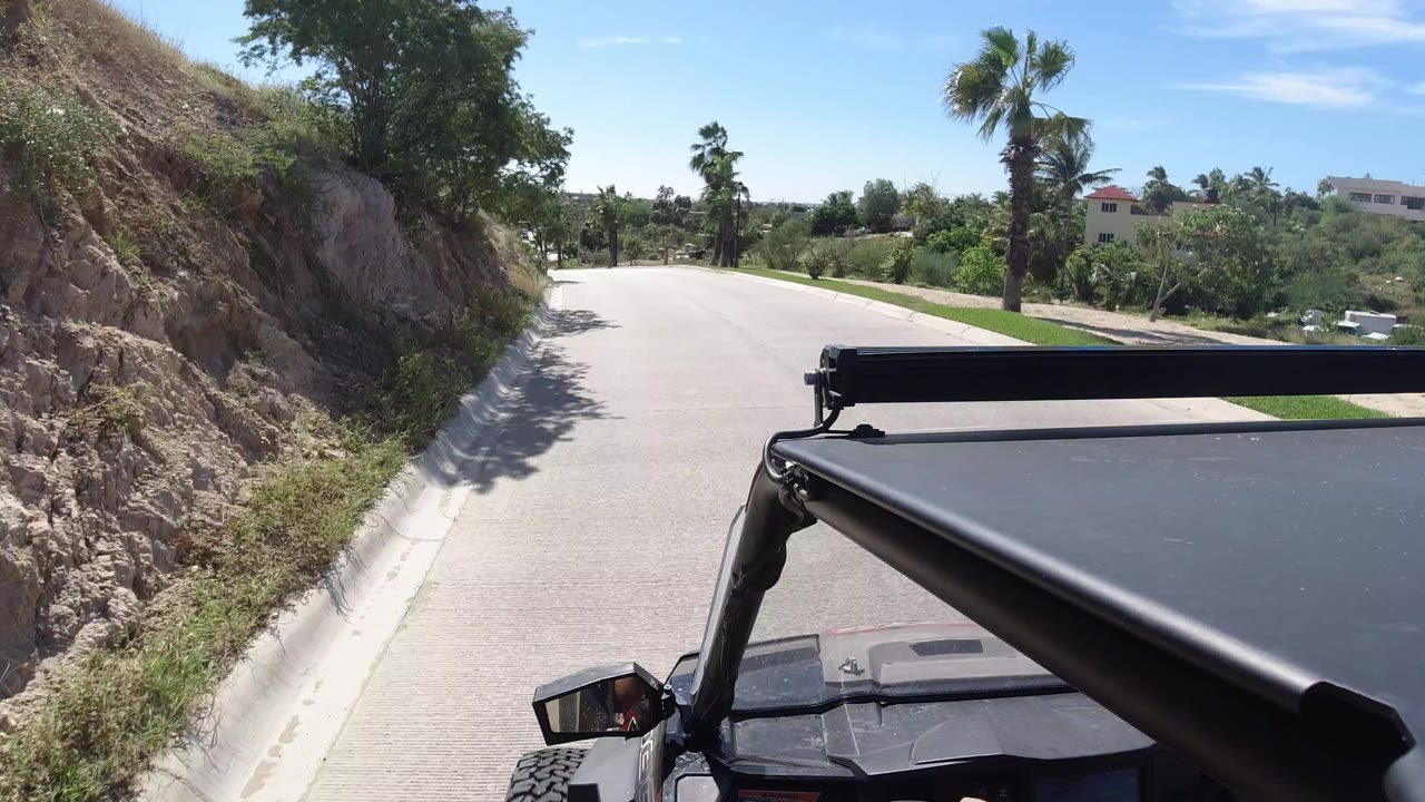A similar Los Cabos road view shows the vehicle roof lower right, the road bending left, dry rock and brush on the left shoulder, palms ahead, distant homes, and no visible people.
