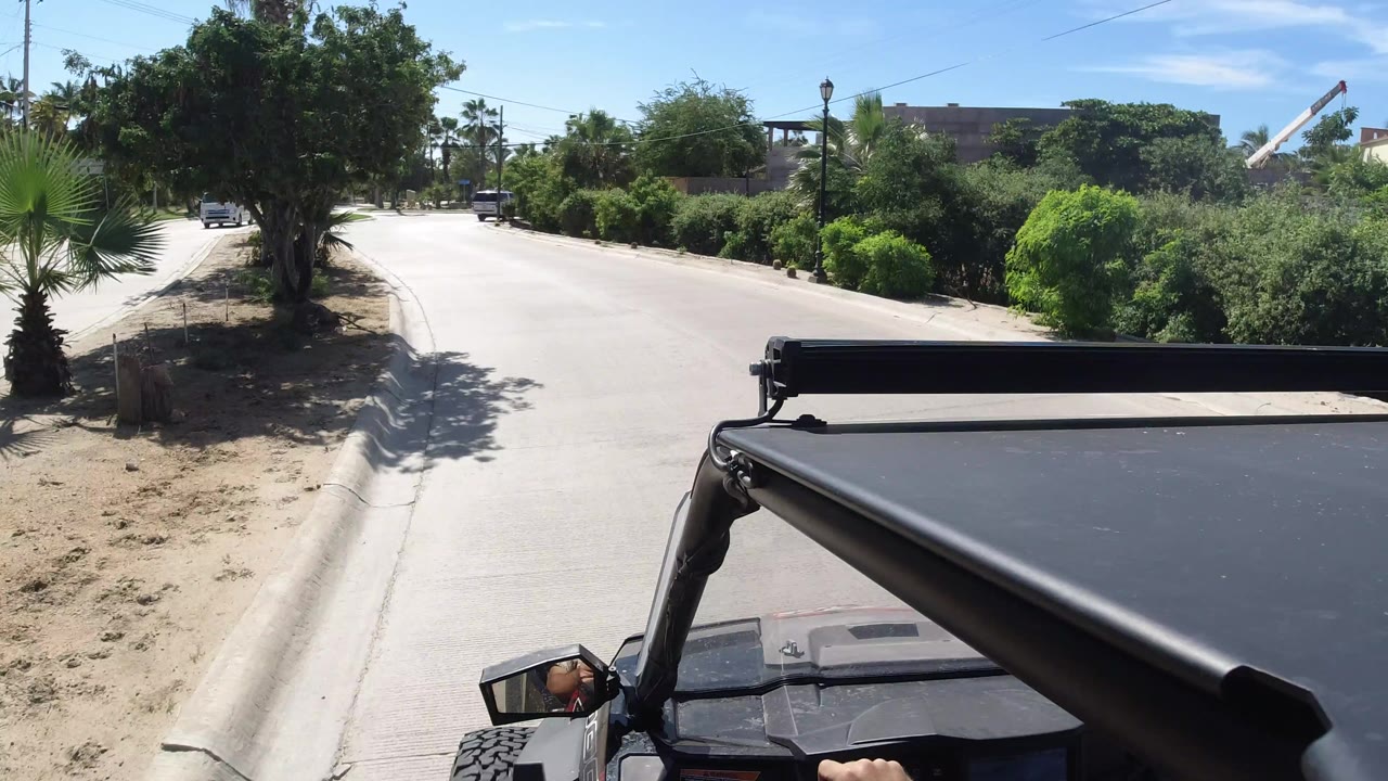 The view continues along the Los Cabos roadside, with the vehicle roof bar filling the lower right, a broad paved lane ahead, dense green shrubs, palms, and a low building beyond them.
