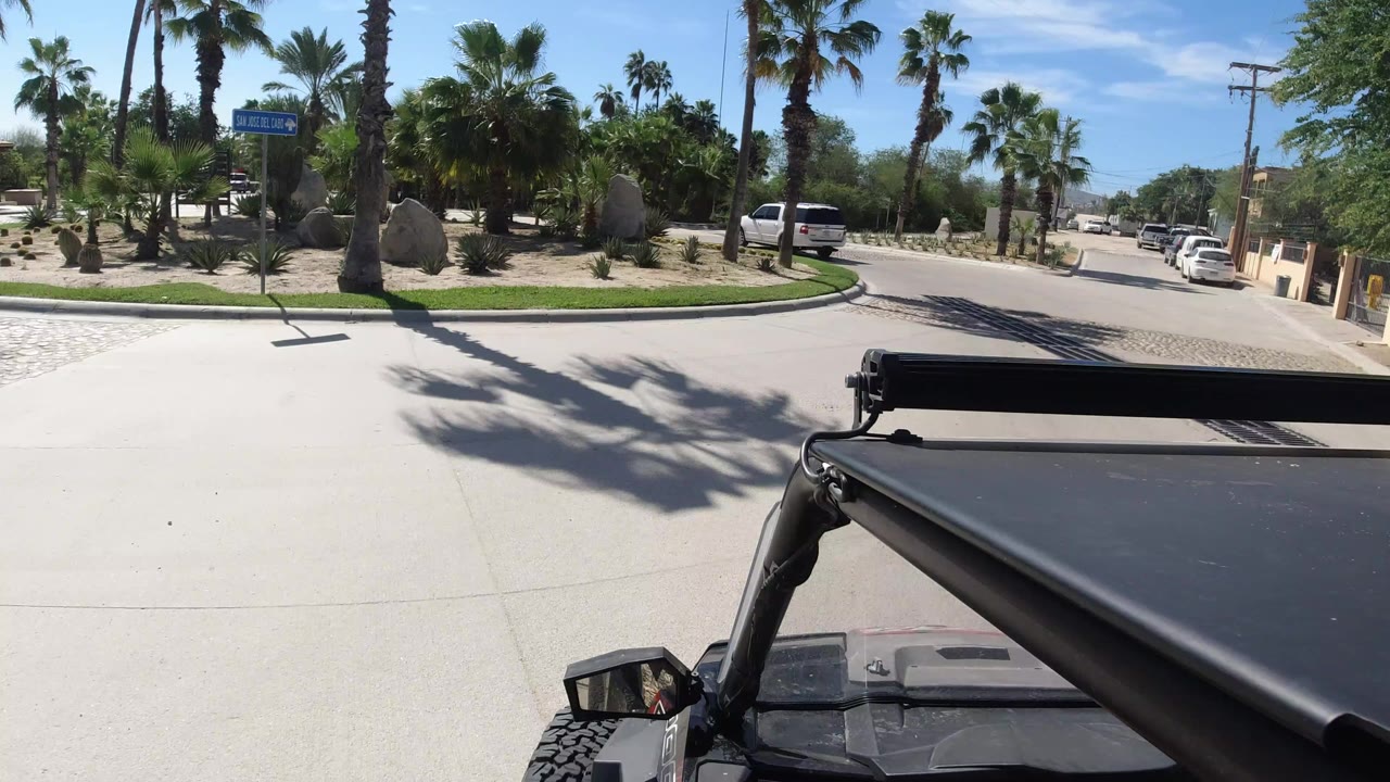 A Los Cabos road scene seen over a black Wrangler roof and side mirror, with palms, desert landscaping, parked vehicles, and low buildings under bright sun, with no clearly visible people.
