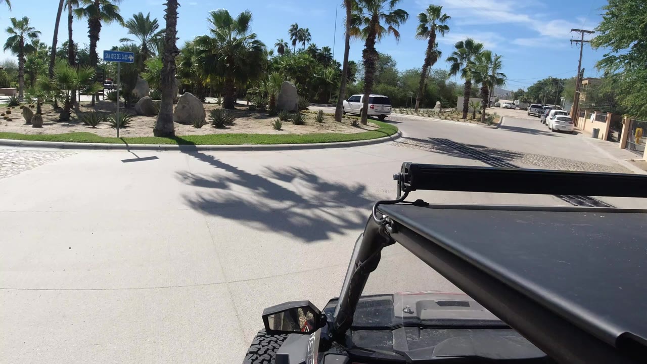 The Wrangler foreground remains steady as the road bends ahead, with a white parked vehicle, palm trees, desert boulders, and a small roadside sign visible in Los Cabos, with no clearly visible people.
