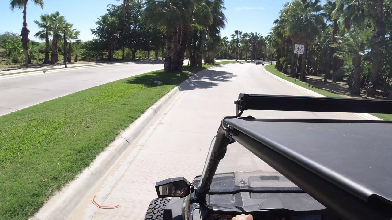 The vehicle moves along the curbside lane in Los Cabos, Mexico, with a green median, palms on both sides, distant traffic, and no clearly recognizable people.
