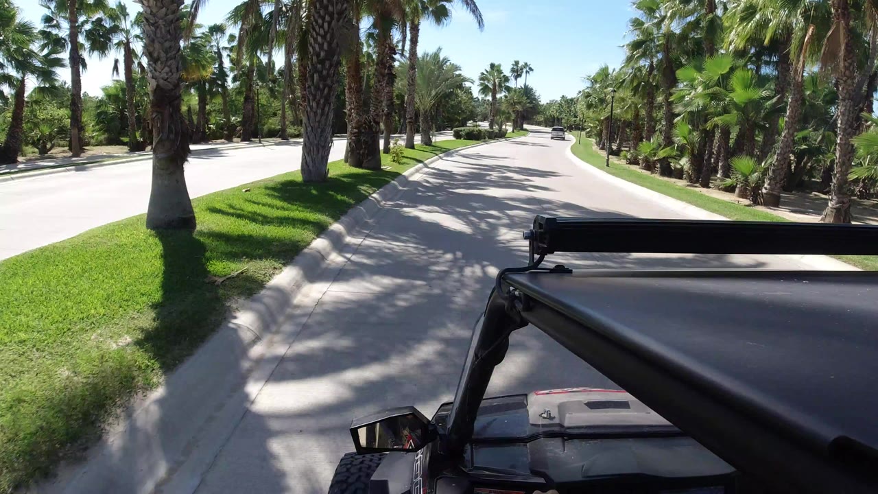 Opening frame of a Los Cabos road view from a black Jeep, with palms on both sides, bright grass along the curb, blue sky overhead, and no visible people.