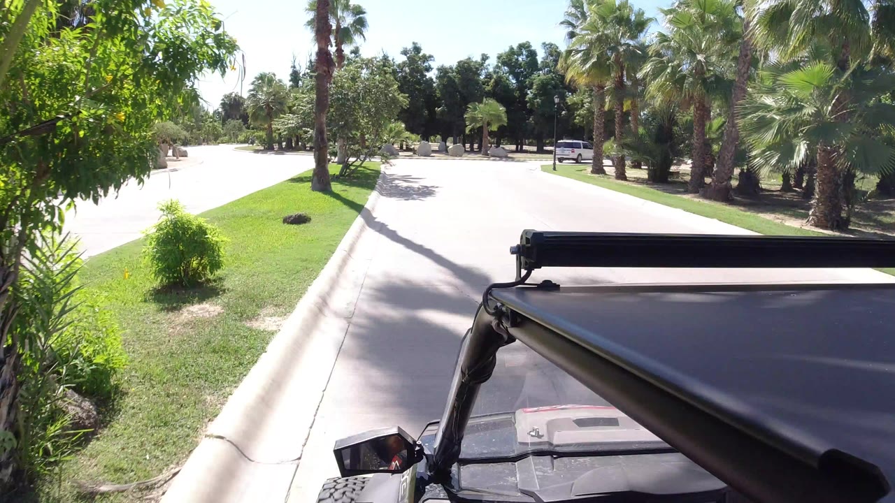 Forward view from a Wrangler in Los Cabos, Mexico, with the roof and mirror in the foreground, a pale paved road ahead, trimmed grass, palms, shrubs, and no clearly visible people.