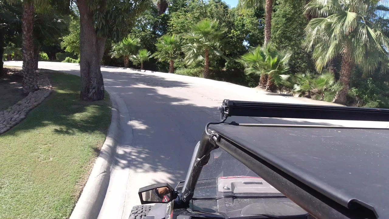 The Wrangler foreground remains fixed while the road bends past a grass edge and palms in Los Cabos, Mexico, with no visible people.