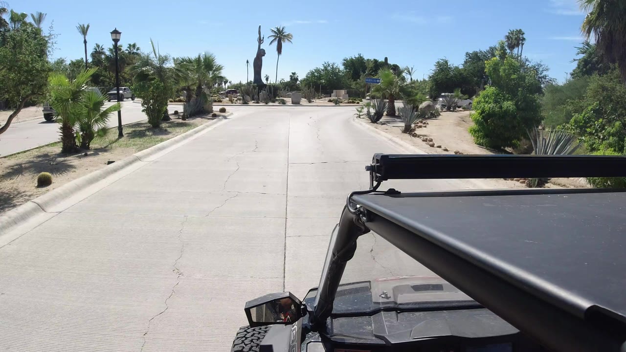 The vehicle continues along the road in Los Cabos, Mexico, with the black foreground frame, bright pavement, palms, shrubs, and a clear blue sky, no visible people.