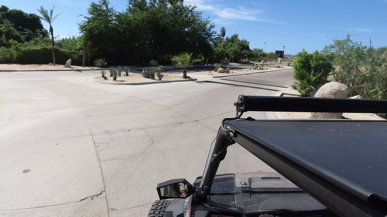 Opening frame of a Wrangler viewpoint in Los Cabos, Mexico, facing a paved road bordered by sand, rocks, palms, and dense green vegetation, with no visible people.